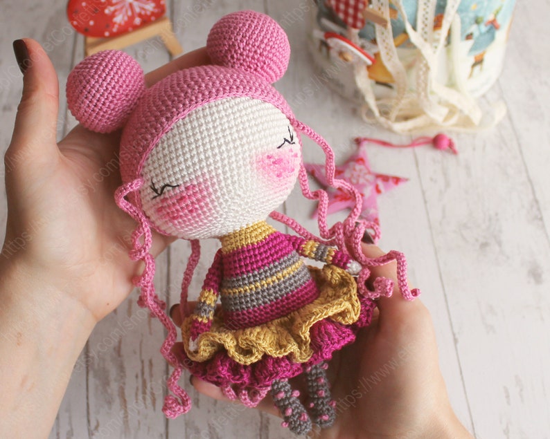 Pu&ograve; includere: Una bambola all'uncinetto con capelli rosa, un cappello rosa e bianco, un vestito a righe rosa e grigio e una gonna a balze gialla.