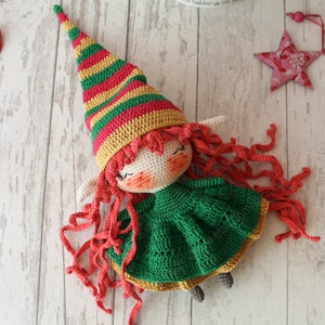 Peut inclure: Une poup&eacute;e elfe en crochet avec de longs cheveux roux, une robe verte et un chapeau ray&eacute; rouge, jaune et vert.