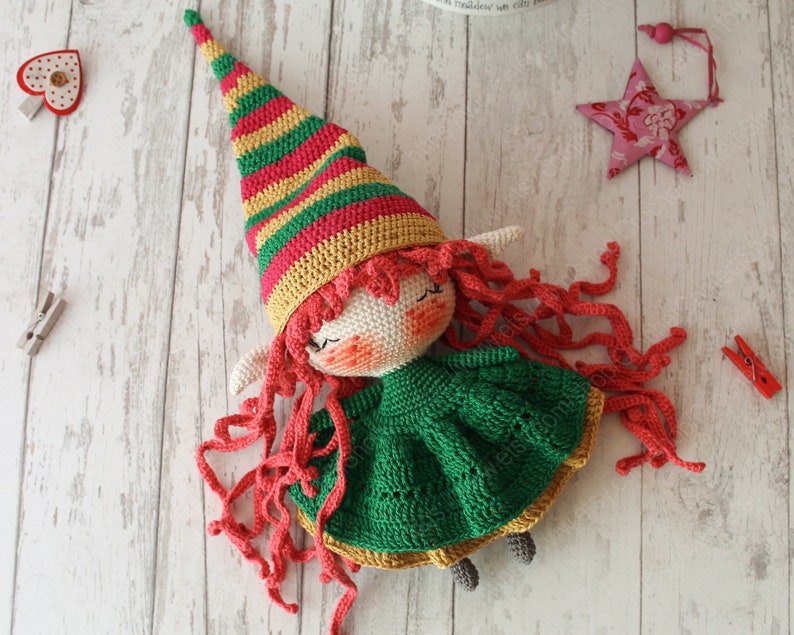Peut inclure: Une poup&eacute;e elfe en crochet avec de longs cheveux roux, une robe verte et un chapeau ray&eacute; rouge, jaune et vert.