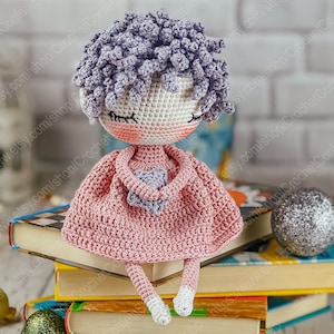 May include: A crocheted doll with purple hair, wearing a pink dress and holding a purple heart. The doll is sitting on a stack of books.