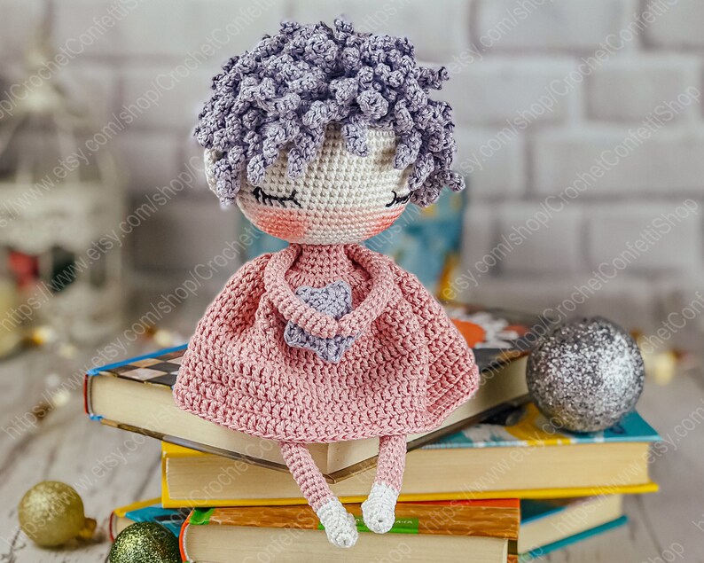 May include: A crocheted doll with purple hair, wearing a pink dress and holding a purple heart. The doll is sitting on a stack of books.