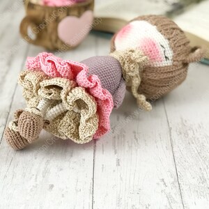 May include: A crocheted doll with brown hair, a purple dress, and a pink and beige ruffled skirt. The doll is lying on a white wooden surface.