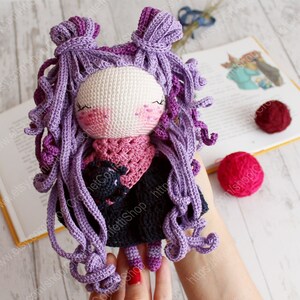 K&ouml;nnte beinhalten: H&auml;kelpuppe mit lila lockigem Haar, die einen rosa Schal und ein schwarzes Kleid tr&auml;gt. Die Puppe h&auml;lt eine kleine schwarze geh&auml;kelte Katze.