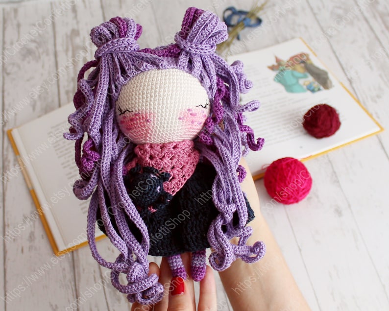 K&ouml;nnte beinhalten: H&auml;kelpuppe mit lila lockigem Haar, die einen rosa Schal und ein schwarzes Kleid tr&auml;gt. Die Puppe h&auml;lt eine kleine schwarze geh&auml;kelte Katze.