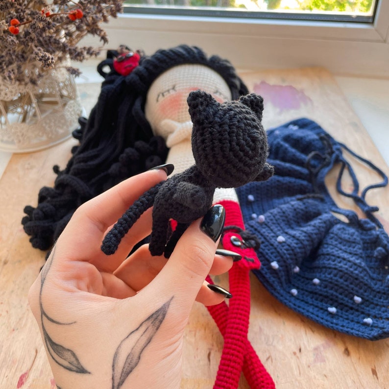 K&ouml;nnte beinhalten: Eine geh&auml;kelte Puppe mit schwarzen Haaren und einem roten Kleid, die eine schwarze geh&auml;kelte Katze h&auml;lt.