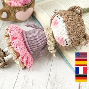 May include: Crochet amigurumi doll pattern in English, Spanish, French, and German. The doll is brown with a pink and beige dress and is lying on a book. The doll has a sweet expression with closed eyes.