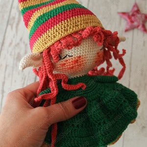 Peut inclure: Une poup&eacute;e elfe en crochet avec des cheveux roux, une robe verte et un chapeau ray&eacute;. La poup&eacute;e est tenue par une main.