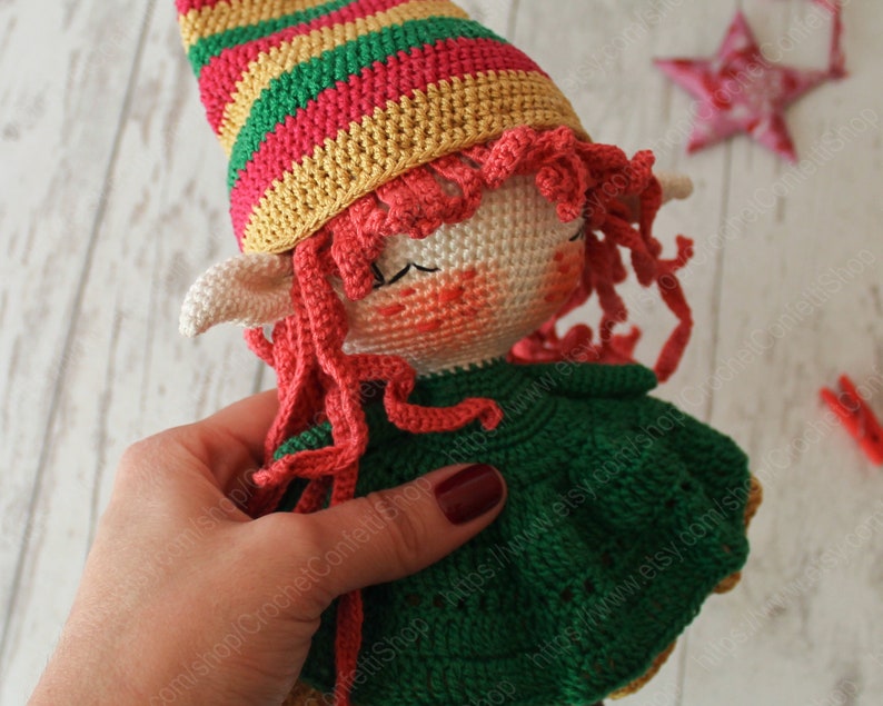 Peut inclure: Une poup&eacute;e elfe en crochet avec des cheveux roux, une robe verte et un chapeau ray&eacute;. La poup&eacute;e est tenue par une main.