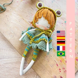 May include: Crocheted doll with a green and blue dress, orange hair, and a frog-shaped hat. The doll is sitting on a wooden surface.