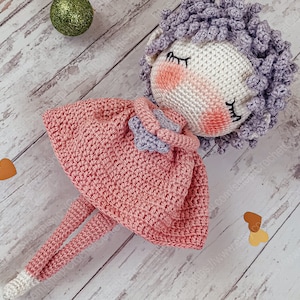 May include: A crocheted doll with pink and purple yarn, wearing a pink dress and holding a heart. The doll is lying on a white wooden surface with brown hearts.