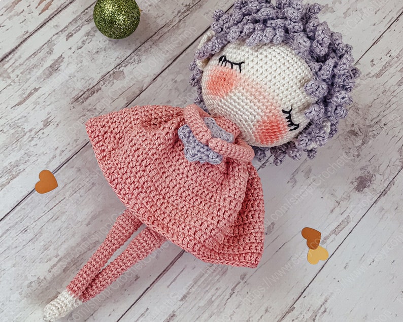 May include: A crocheted doll with pink and purple yarn, wearing a pink dress and holding a heart. The doll is lying on a white wooden surface with brown hearts.