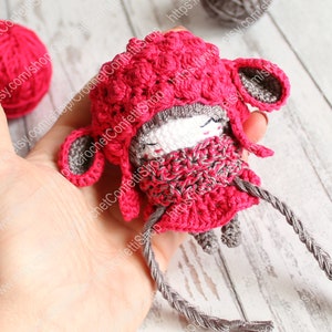 May include: A small crocheted doll wearing a pink hat and a gray scarf. The doll has a gray face with black eyes and a pink nose. The doll is holding a gray string in its hands.