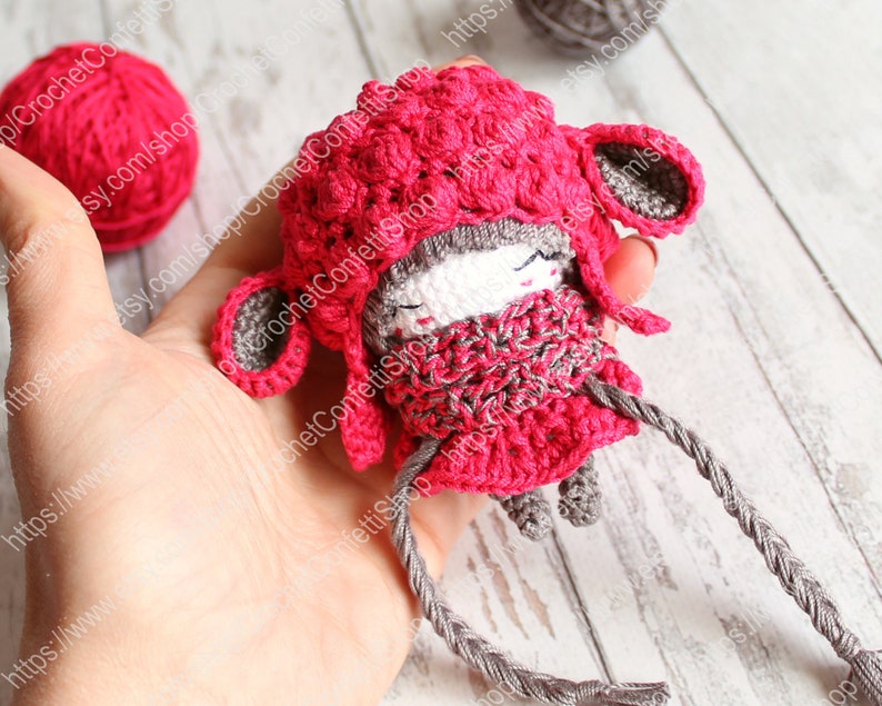 May include: A small crocheted doll wearing a pink hat and a gray scarf. The doll has a gray face with black eyes and a pink nose. The doll is holding a gray string in its hands.