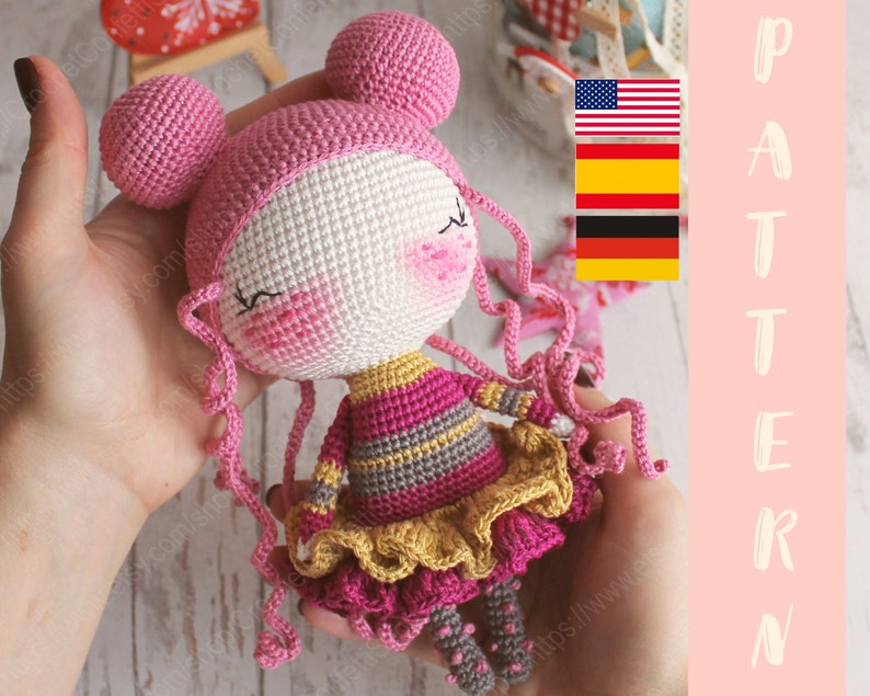 Pu&ograve; includere: Una bambola all'uncinetto con capelli rosa, viso bianco e vestito a righe rosa e grigio. La bambola tiene un fiore giallo in mano. La bambola &egrave; seduta su una superficie di legno. L'immagine include anche la parola "PATTERN" in grandi lettere.