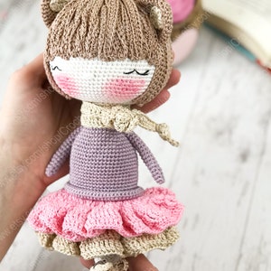 May include: A crocheted doll with brown hair, a pink dress, and a purple sweater. The doll is wearing a cream-colored scarf and has a sweet expression on its face.