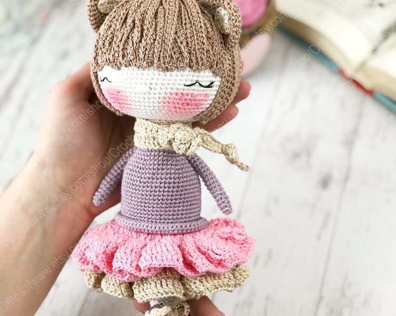 May include: A crocheted doll with brown hair, a pink dress, and a purple sweater. The doll is wearing a cream-colored scarf and has a sweet expression on its face.