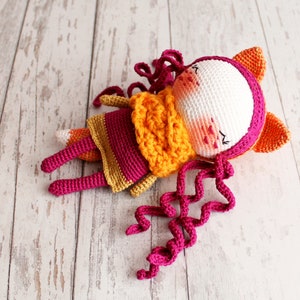 May include: A handmade crochet fox doll in a lying position. The doll features a white face with pink cheeks, a magenta hood, and orange ears. It wears a yellow and magenta dress with curly magenta hair. The fox has orange and magenta legs and a fox tail.
