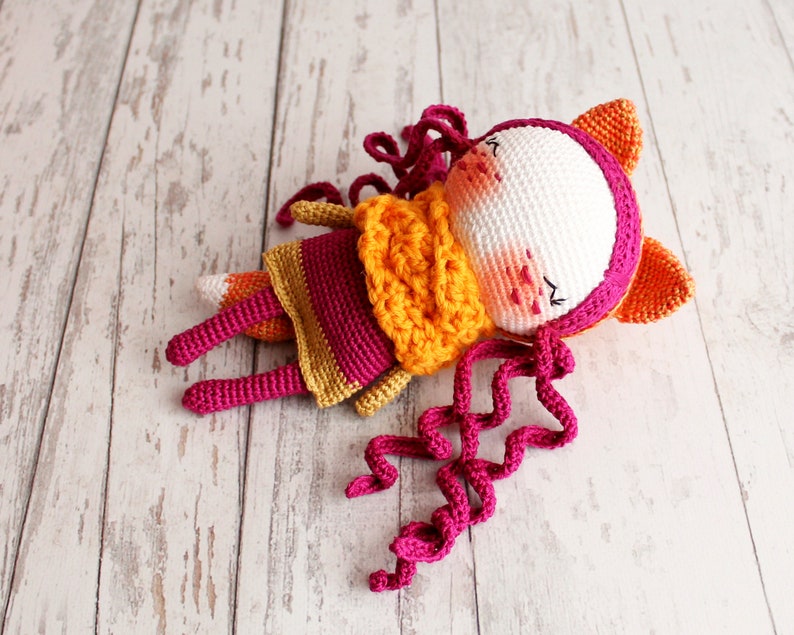 May include: A handmade crochet fox doll in a lying position. The doll features a white face with pink cheeks, a magenta hood, and orange ears. It wears a yellow and magenta dress with curly magenta hair. The fox has orange and magenta legs and a fox tail.