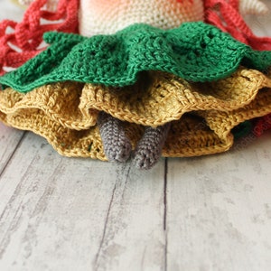 Peut inclure: Une poup&eacute;e en crochet avec une robe verte, jaune et orange. La poup&eacute;e a des cheveux en laine rouge et des jambes en crochet grises.