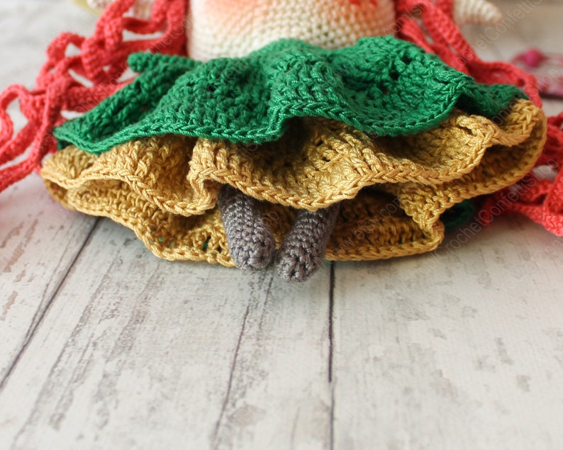 Peut inclure: Une poup&eacute;e en crochet avec une robe verte, jaune et orange. La poup&eacute;e a des cheveux en laine rouge et des jambes en crochet grises.