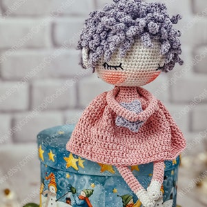 May include: A crocheted doll with purple hair, wearing a pink dress and holding a purple star. The doll is sitting on a blue and white box with a winter scene.
