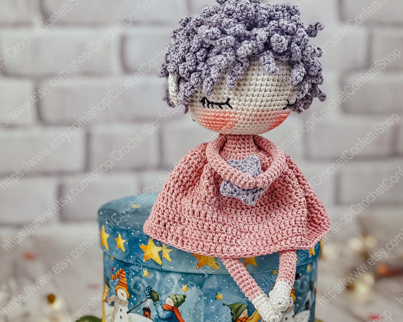 May include: A crocheted doll with purple hair, wearing a pink dress and holding a purple star. The doll is sitting on a blue and white box with a winter scene.