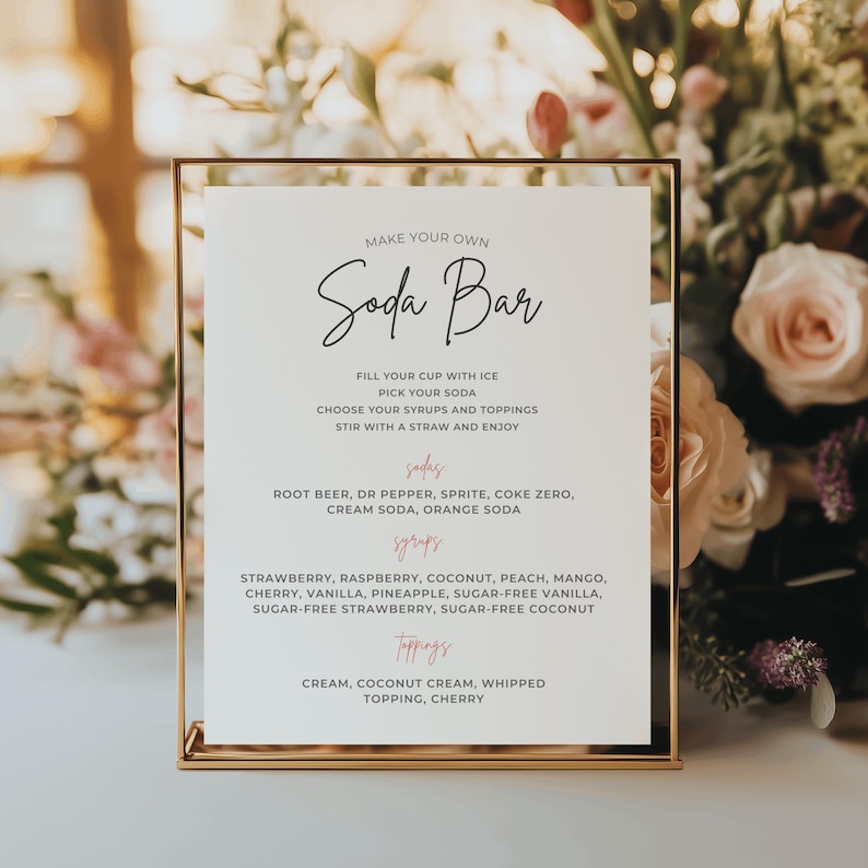 Może przedstawiać: Złota ramka z napisem "MAKE YOUR OWN Soda Bar" w eleganckiej czcionce. Znak wymienia smaki sody, syropy i dodatki. Tło przedstawia miękkie kwiaty, tworząc dekoracyjną ekspozycję na imprezę lub wydarzenie.