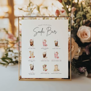 May include: A gold framed sign with the text "MAKE YOUR OWN Soda Bar" in elegant script. The sign features watercolor illustrations of various soda concoctions, including cola, pink fizz, and cream soda, with ingredients listed below each.