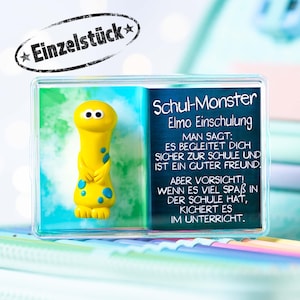 Könnte beinhalten: Eine gelbe Monsterfigur mit blauen Punkten, in einer durchsichtigen Plastikbox mit weißem Hintergrund. Der Text auf der Box lautet "Schul-Monster Elmo Einschulung" und enthält eine kurze Botschaft darüber, dass das Monster ein guter Freund ist.