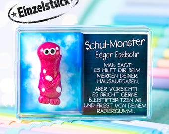 May include: A clear plastic case containing a small, glittery magenta monster figurine with white spots and a card featuring the text Schul-Monster Edgar Eselsohr, Man sagt: Es hilft dir beim Merken deiner Hausaufgaben. Aber Vorsicht! Es bricht gerne Bleistiftspitzen ab und frisst von deinem Radiergummi. The top of the case is labeled Einzelstück.