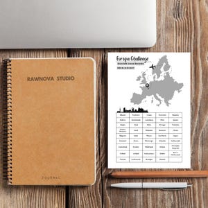 Könnte beinhalten: Ein braunes Spiralnotizbuch mit dem Aufdruck "RAWNOVA STUDIO" und "JOURNAL" auf dem Einband, neben einer gedruckten Europakarte mit dem Thema Reise-Challenge. Ein Stift und ein Bleistift liegen ebenfalls auf der Holzoberfläche.