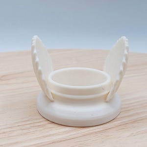 Könnte beinhalten: Ein cremefarbener Eierbecher mit rundem Sockel und zentralem Becher. Zwei flügelförmige Elemente erstrecken sich von den Seiten nach oben. Der Eierbecher besteht aus einem glatten, festen Material.