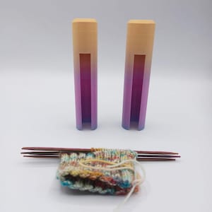 Könnte beinhalten: Zwei zylindrische Stricknadelhalter mit einem Farbverlauf von Pfirsich, Lila und Blau. Ein kleines Stück Strickstoff wird von Stricknadeln aus Holz gehalten. Der Stoff hat ein farbenfrohes, strukturiertes Muster.