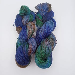 Könnte beinhalten: Zwei Stränge handgefärbter Wolle in Blau-, Grün- und Brauntönen. Das Garn ist zu einer lockeren, strukturierten Form verdreht, wobei die Farben ineinander übergehen. Das Garn wirkt weich und eignet sich für Strick- oder Häkelprojekte.