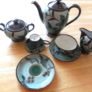 Op de afbeelding: Een keramische theeset met een theepot, suikerpot, melkkan, twee kopjes en schotels. De set heeft een teal en donkergrijs kleurenschema met een botanisch ontwerp van bladeren en bessen. De theeset staat op een houten oppervlak.