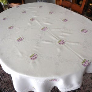 Peut inclure: Une nappe ovale blanche présentant un motif damassé ton sur ton subtil et des grappes de fleurs violettes brodées avec des feuilles vertes. Les bords du tissu sont finis avec une délicate bordure en dentelle blanche sur tout le pourtour.