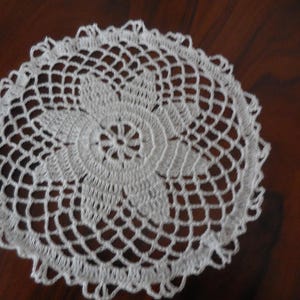Può includere: Tovaglietta all'uncinetto bianca con un delicato disegno floreale. La tovaglietta è circolare e ha un bordo in pizzo.