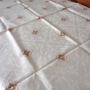 Könnte beinhalten: Eine weiße Tischdecke mit einem dezenten Ton-in-Ton-Damastmuster auf dem Stoff. Die Oberfläche ist mit gleichmäßig verteilten, kleinen quadratischen Kreuzstich-Stickmotiven in den Farben Rot, Grün und Braun über das gesamte Design hinweg verziert.