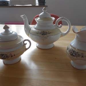 Può includere: Un servizio da tè vintage con teiera, zuccheriera e lattiera. I pezzi in porcellana bianca presentano una fascia giallo pallido con decorazioni floreali e bordi dorati. La teiera ha un beccuccio curvo e un manico arrotondato.