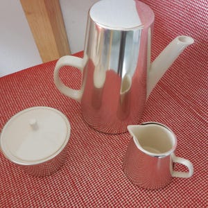 Op de afbeelding: Een driedelige koffieset bestaande uit een hoge koffiepot, een suikerpot met deksel en een kleine melkkan. Elk onderdeel heeft een glanzende, zilverkleurige metalen behuizing met contrasterende crèmekleurige keramische handvatten, deksels en tuiten.