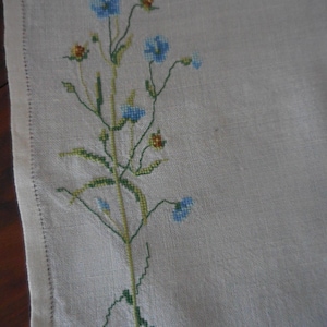 Könnte beinhalten: Nahaufnahme eines weißen Leinentextils mit einem Kreuzstich-Blumendesign. Die Stickerei zeigt blaue Blumen, grüne Stiele und kleine orangefarbene Akzente. Der Rand des Textils hat eine genähte Bordüre.