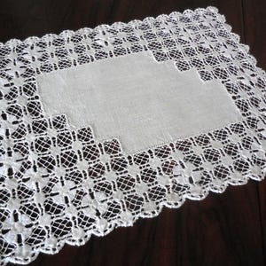May include: A white rectangular doily with a central plain area and an intricate lace border. The lace features a repeating geometric pattern with scalloped edges. The doily is placed on a dark wooden surface.