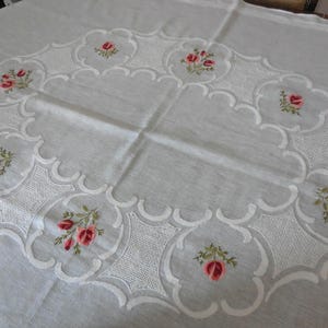 Peut inclure: Une nappe rectangulaire blanche ornée d'une bordure ovale centrale composée de bords festonnés et de broderies blanches complexes. Le motif est agrémenté de petits motifs floraux colorés de roses roses et de feuilles vertes le long du bord.