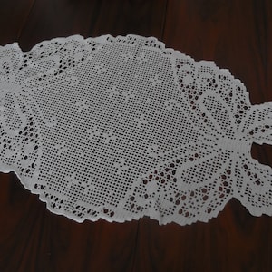 Könnte beinhalten: Weißer gehäkelter Tischläufer mit einem zarten Blumenmuster. Der Läufer ist oval geformt und hat einen Spitzenrand.