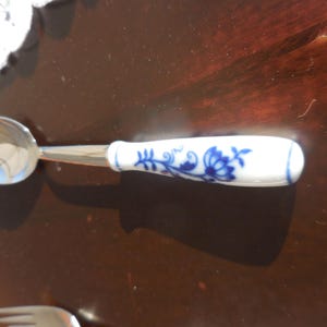 Può includere: Un cucchiaino con una ciotola lucida color argento e un manico bianco decorato con motivi floreali blu. Il cucchiaino è probabilmente per servire o mangiare, e il design del manico aggiunge un tocco di eleganza.