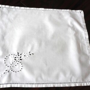 May include: A square white napkin featuring a delicate cutwork embroidery pattern in one corner. The design consists of circular motifs and small floral elements. The edges of the napkin are finished with a decorative hemstitch border detail.