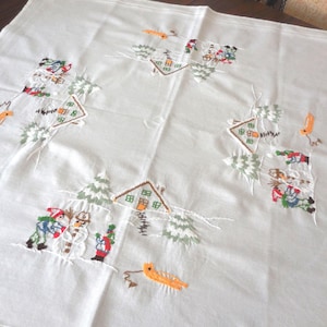 Può includere: Una tovaglia quadrata bianca con scene invernali ricamate disposte in un motivo circolare. Il design include piccole case, alberi sempreverdi, pupazzi di neve, figure con abiti invernali colorati e slitte arancioni cucite sul tessuto.