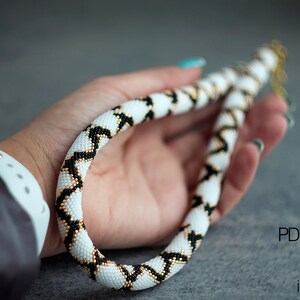 May include: A white, black and gold beaded necklace with a geometric pattern. The necklace is held in a hand and is approximately 46 cm long. PDF pattern MRF jewellery.