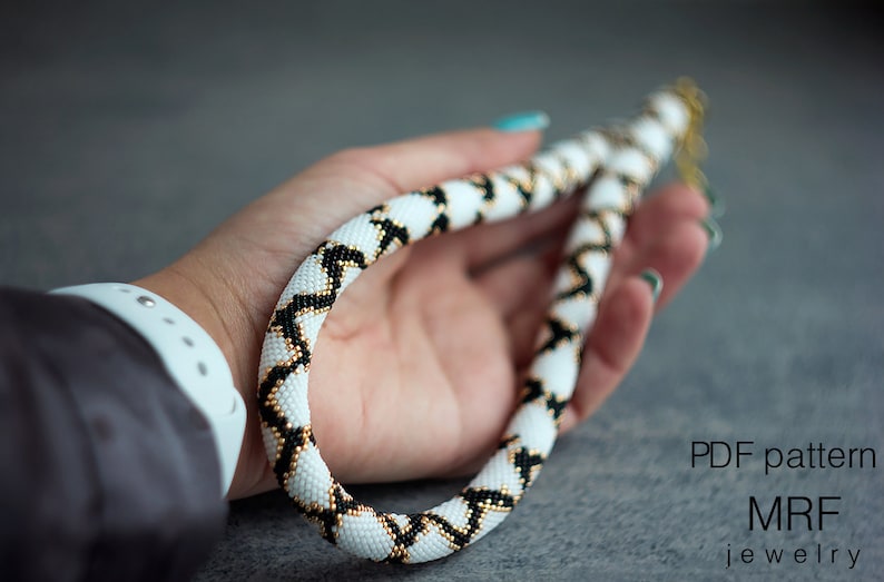 May include: A white, black and gold beaded necklace with a geometric pattern. The necklace is held in a hand and is approximately 46 cm long. PDF pattern MRF jewellery.