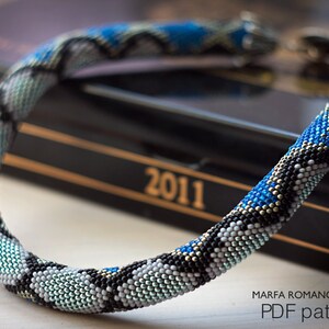Może przedstawiać: Naszyjnik z koralik&oacute;w w kształcie sk&oacute;ry węża w odcieniach niebieskiego, czarnego, białego i srebrnego. Naszyjnik leży na czarnej książce z nadrukowanym rokiem 2011 na okładce. Pod naszyjnikiem znajduje się napis "MARFA ROMANOVSKAYA PDF pattern".